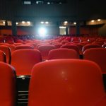Sala de cinema vazia, com fileiras de poltronas vermelhas voltadas para uma tela iluminada.