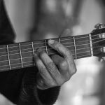Mãos de um músico tocando violão acústico em uma fotografia em preto e branco.