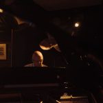 Pianista idoso toca piano em um ambiente escuro, com iluminação intimista.