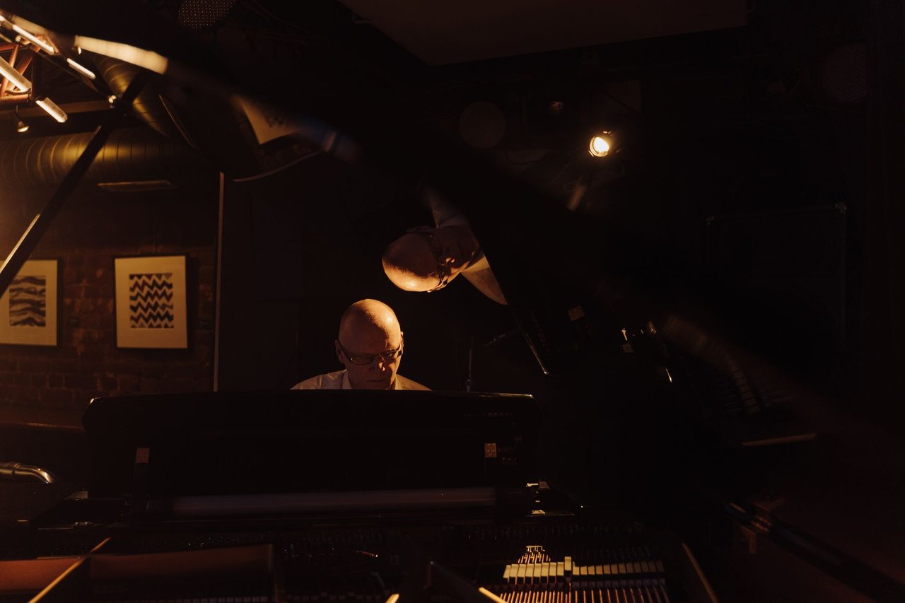 Pianista idoso toca piano em um ambiente escuro, com iluminação intimista.