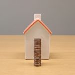 Moedas empilhadas diante de uma pequena casa de porcelana, representando economia e investimento imobiliário.