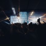 Público acompanha um show noturno ao ar livre, diante de um palco iluminado por luzes coloridas.